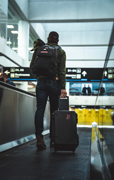 Voyageur à l'aéroport avec une mallette souple SKB noire à roulettes et un sac à dos, adaptés au transport sécurisé d'équipements lors de voyages.