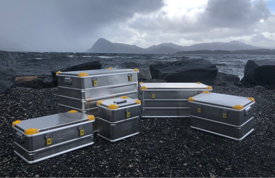 Boîtes de transport Viking en aluminium avec coins jaunes disposées sur une côte rocheuse, avec la mer et un paysage montagneux en arrière-plan.