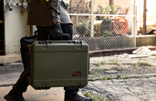 Valise rigide SKB portée par une personne dans un environnement industriel avec sol en béton et clôture en arrière-plan.