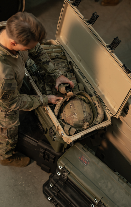 Valise rigide SKB ouverte, dans laquelle est rangé du matériel militaire, tandis qu'une personne en uniforme militaire range le contenu dans un environnement intérieur.