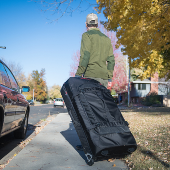 Sac en nylon SKB à roulettes, tiré par une personne sur un trottoir dans un quartier résidentiel avec des voitures garées et des arbres en automne.