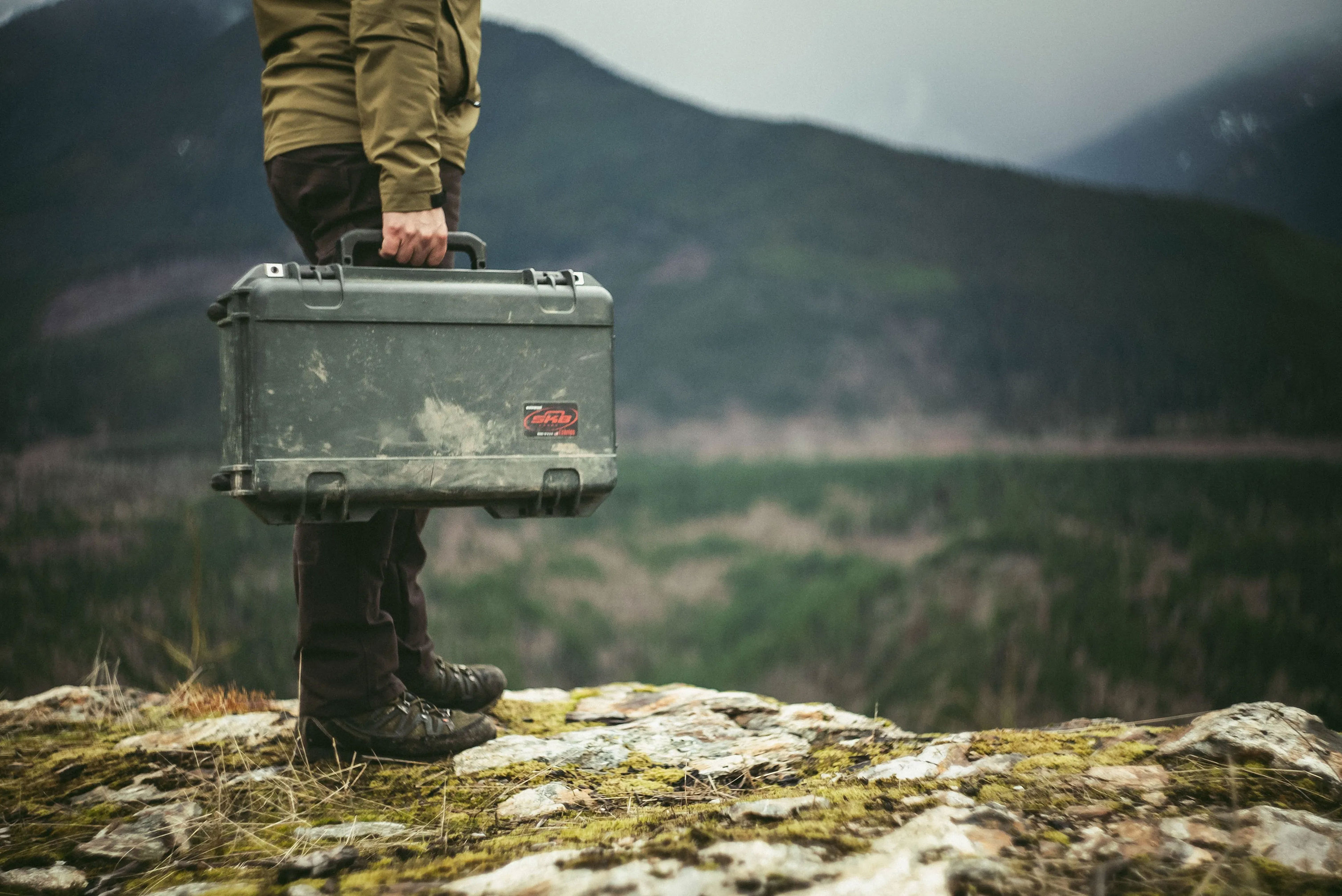 Valise rigide SKB transportée par une personne sur un terrain rocheux dans un environnement montagneux.